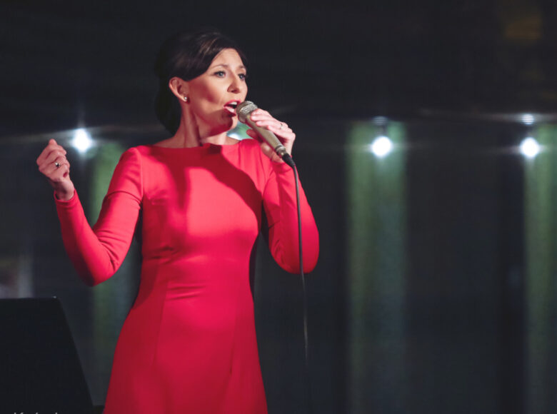 Zdjęcie. Elegancko uczesana kobieta w długiej, czerwonej sukni z długimi rękawami stoi na scenie i śpiewa.