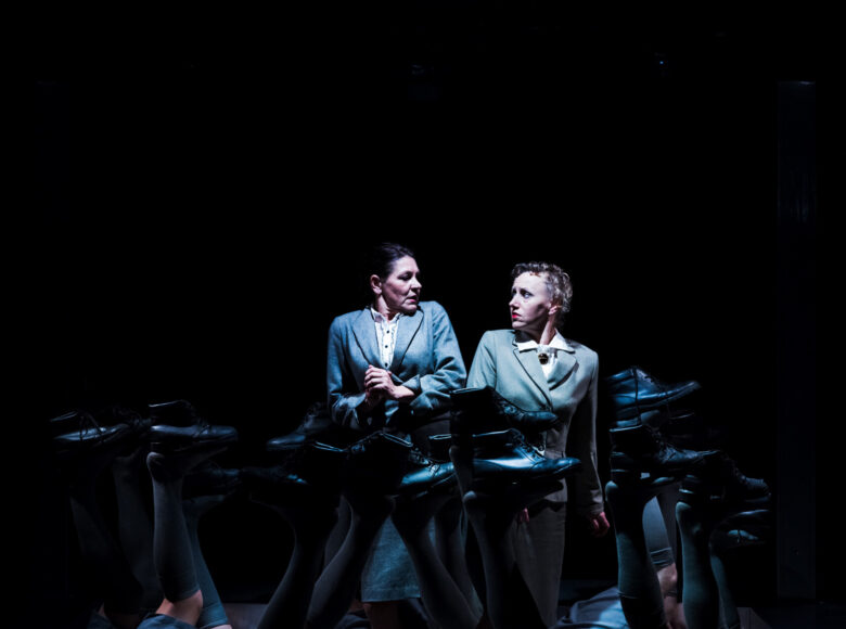 Zdjęcie. Scena ze spektaklu "Chór sierot". Na środku sceny stoją dwie kobiety w garsonkach. Patrzą na siebie z przestrachem. Kobiety otacza grupa ludzi, leżących na podłodze i unoszących do góry nogi, na stopach podtrzymują po jednym czarnym bucie.
