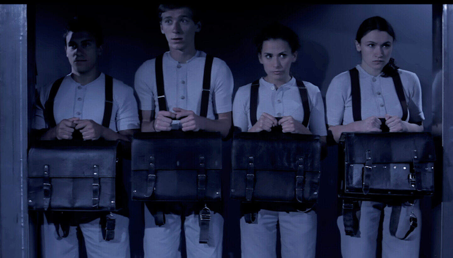 Zdjęcie. Scena ze spektaklu "Chór sierot". W równym rzędzie stoi grupa młodych ludzi, chłopców i dziewczyn. Są identycznie ubrani: w białe koszulki i szare spodnie z czarnymi szelkami. Przed sobą trzymają tornistry. Sprawiają wrażenie smutnych lub przestraszonych.