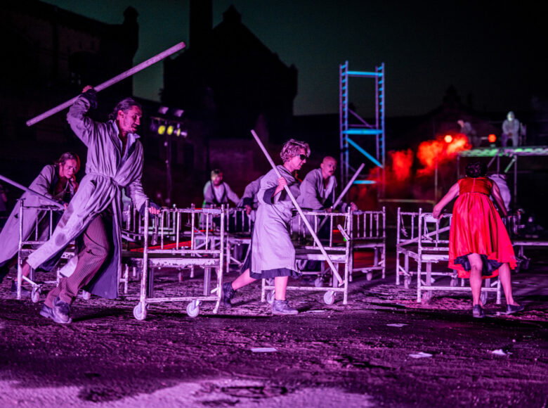 Zdjęcie. Scena ze spektaklu. Grupa ludzi w szpitalnych strojach i z laskami w rękach biegnie po kole. Przed sobą pchają szpitalne łózka na kółkach.