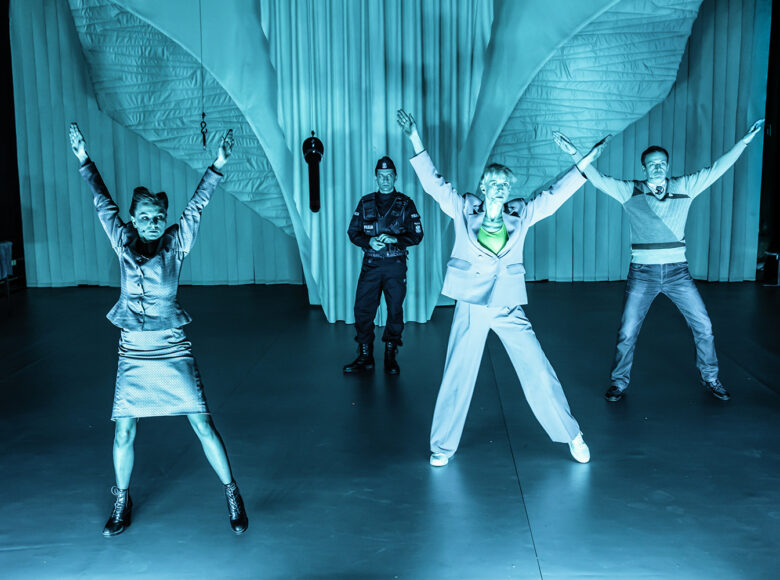 Na zdjęciu scena grupowa. Dwie kobiety i mężczyzna stoją w takich samych pozach – nogi szeroko rozstawione, ręce uniesione nad siebie, pozycje przypominają literę X. Z tyłu za nimi stoi mężczyzna w mundurze policyjnym. Scena oświetlona szmaragdowym światłem.