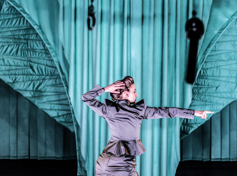 Kobieta ubrana w srebrny, elegancki komplet, stojąca plecami do obiektywu. Jedną rękę ma wyciągniętą w bok, twarz także zwrócona w tę stronę. Drugą rękę trzyma na głowie. W tle szmaragdowozielony, udrapowany materiał.