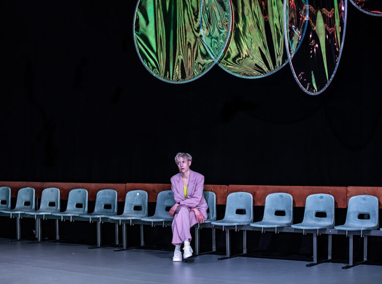 Kobieta siedzi na krześle, jednym w rzędzie krzeseł ustawionych wzdłuż ścianą. Nad nią widoczne, niektóre częściowo, 4 koła z połyskliwym materiałem.