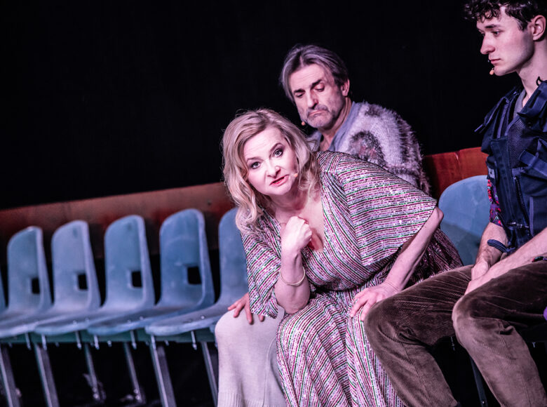 Dwóch mężczyzn i kobieta siedzą na krzesłach. Krzesła ustawione w rzędzie wzdłuż ściany. Kobieta pochyla się do przodu, wydaje się mówić, patrząc w obiektyw.