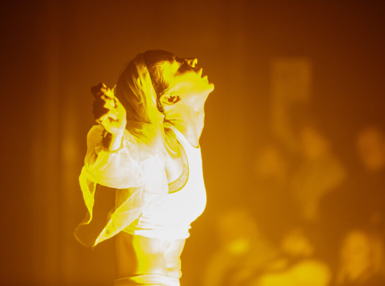 Ujęcie od pasa w górę. Kobieta stoi z uniesionymi na bok rękami. Jej sylwetkę oświetla żółte światło.