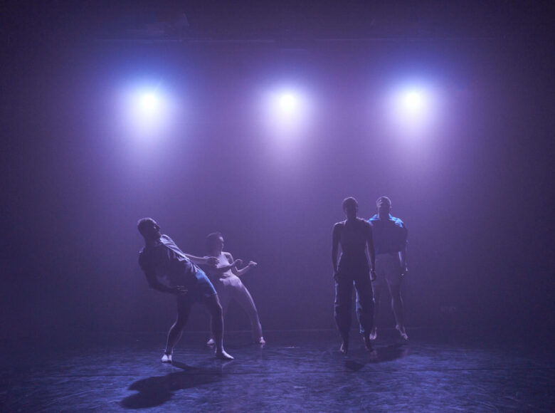 Na scenie stoją cztery osoby. Stoją w parach – jedna za drugą. Osoby po prawej stronie fotografii podskakują, a osoby po lewej stronie odchylają się lekko do tyłu. Nad nimi w tle święcą się trzy reflektory.