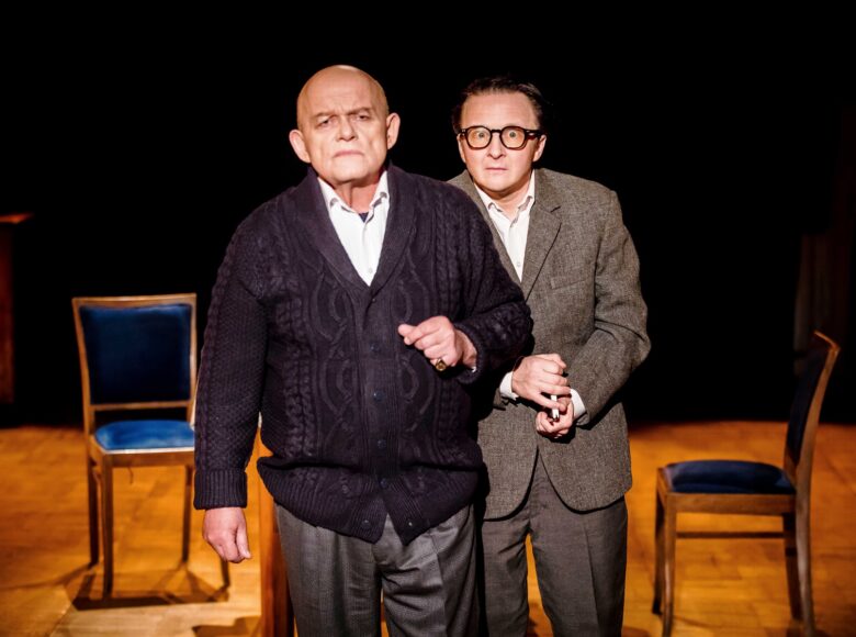 Na pierwszym planie stoi dwóch mężczyzn, jeden łysy, starszy, drugi w okularach. W tle stoją dwa drewniane krzesła.