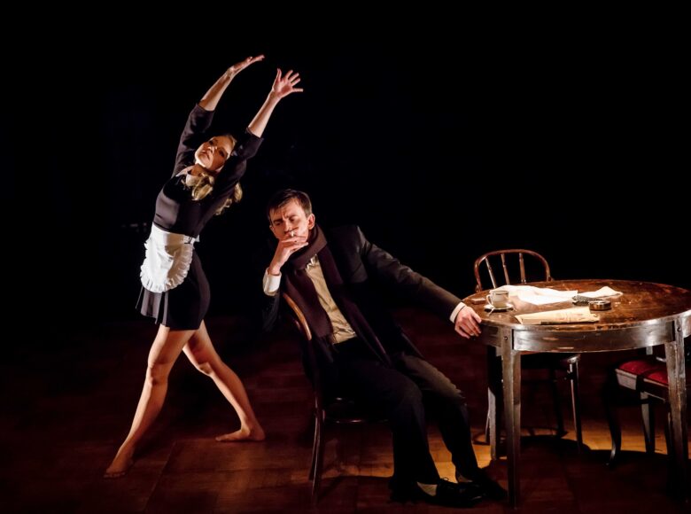 Na zdjęciu dwie osoby, scena częściowo w ciemności. Mężczyzna siedzi w centrum, obok niego okrągły, drewniany stół, na którym stoi filiżanka i leżą rozrzucone kartki papieru. Mężczyzna pali papierosa. Po jego lewej stronie bosa kobieta w stroju kelnerki, z małym białym fartuszkiem. Wydaje się tańczyć, ma ręce uniesione nad głową.