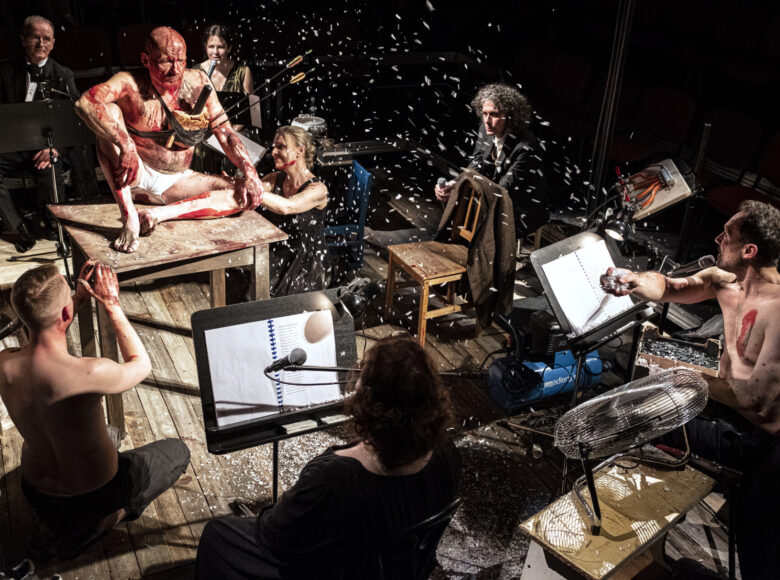 Scena zbiorowa, osoby siedzą na drewnianym podeście. Na środku, na kwadratowym stole siedzi mężczyzna. Ubrany tylko w białe bokserki, na plecach, głowie i nogach widoczne smugi krwi. Wokół stołu kucają 3 osoby, patrząc na mężczyznę. Na pierwszym planie dwie osoby siedzą za pulpitami do nut. Nad całością sceny unosi się pierze.