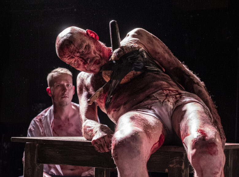 Na zbliżeniu mężczyzna, siedzi na drewnianym stole, opiera się prawą ręką o stół. Jego ciało jest pokryte smugami krwi. Na klatce piersiowej ma doczepiony przedmiot, w który jest wbity nóż. Z tyłu widoczny drugi, siedzący mężczyzna.