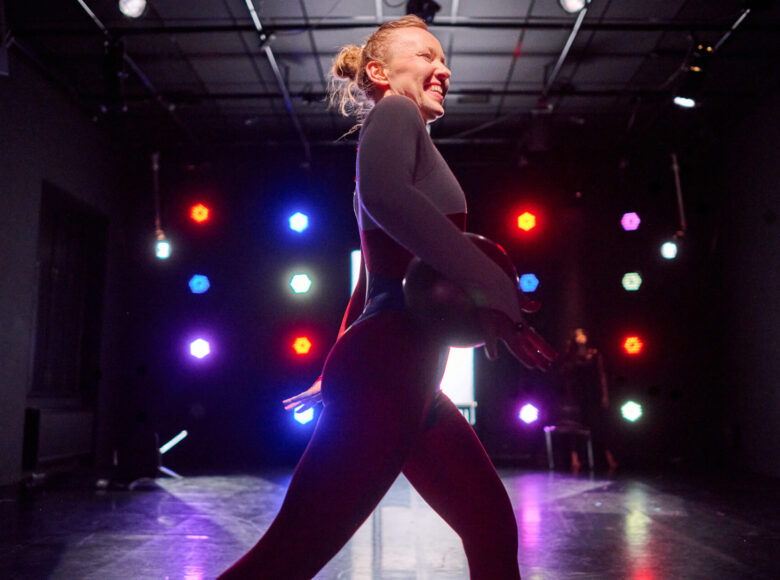 Kolorowa fotografia. Ustawiona bokiem do publiczności młoda kobieta kroczy po scenie. Jest ubrana w obcisły kombinezon a pod pachą trzyma piłkę. W tle scenę oświetlają kolorowe reflektory.