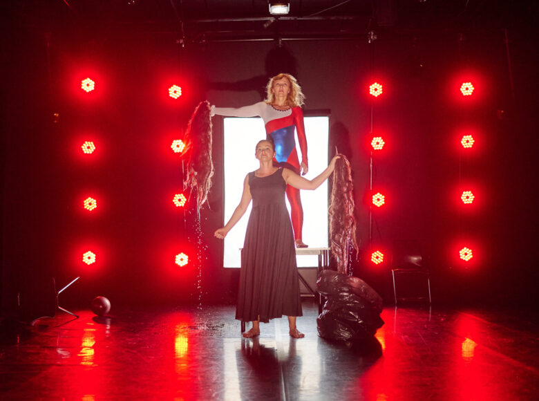 Kolorowa fotografia. Na scenie oświetlonej czerwonymi reflektorami stoją dwie kobiety. Jedna z nich stoi na stoliku i jest ubrana w biało-czerwono-czarny kombinezon, druga w ciemną sukienkę. Kobiety trzymają w ręku peruki zamoczone wodą, która ścieka na podłogę.