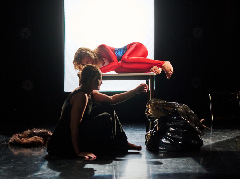 Kolorowa fotografia. Na ciemnej scenie stoi mały stolik, na którym leży bokiem z podkulonymi nogami kobieta, ubrana w obcisły kombinezon. Przed stolikiem na podłodze siedzi druga kobieta. W tle scenę oświetla ekran z białym mocnym światłem.