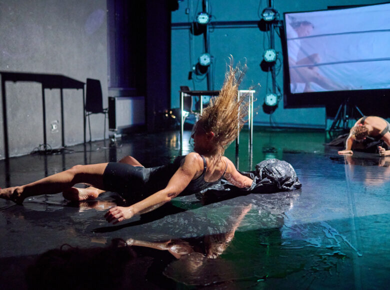 Kolorowa fotografia. Dynamiczna scena przedstawia dwie kobiety tarzające się po mokrej podłodze. Na scenie stoi też stolik oraz widać w tle zawieszony ekran.