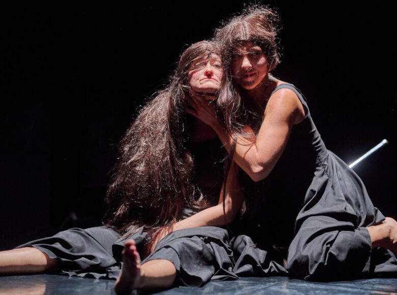 Kolorowa fotografia. Na podłodze sceny siedzą obok siebie dwie kobiety ubrane w ciemne sukienki. Jedna z nich trzyma drugą za kark. Mają na sobie peruki.