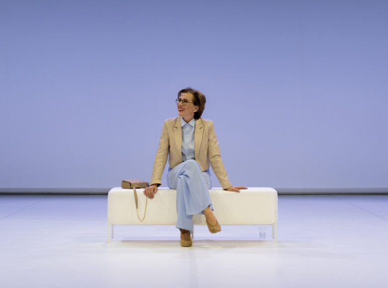 Na środku zdjęcia, na białej, prostokątnej pufie siedzi kobieta, obok niej leży torebka. Kobieta się uśmiecha, patrzy w lewą stronę kadru. Scena skąpana w niebieskim świetle.