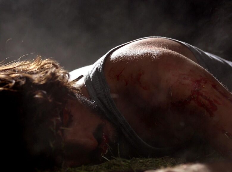 Kadr z filmu. Zbliżenie na głowę i ramiona leżącego na brzuchu, na ziemi, mężczyzny. Jego ramie jest zakrwawione. Silne światło oświetla plecy i kręcące się włosy, twarz z zamkniętymi oczami pozostaje w cieniu.