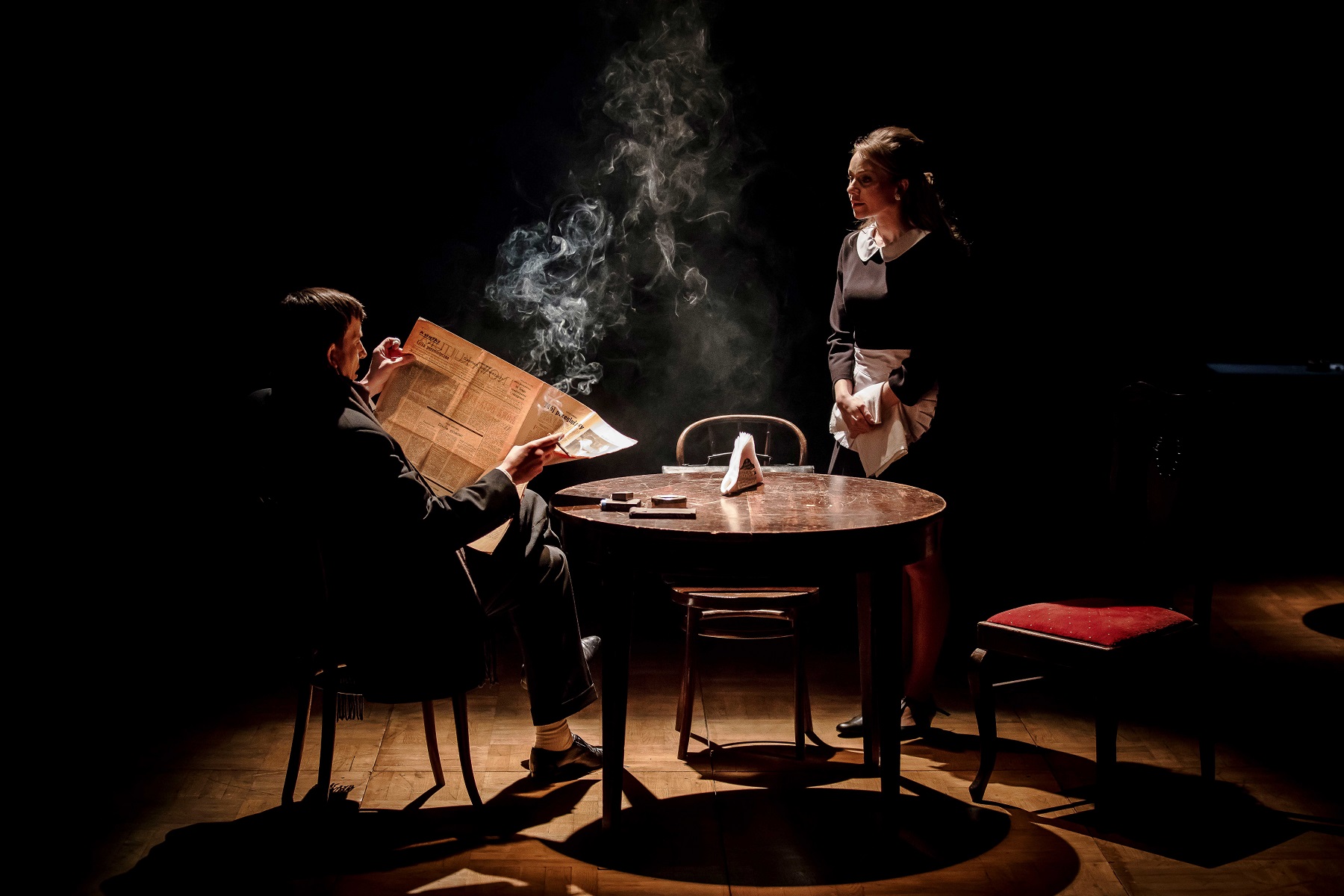 Na zdjęciu dwie osoby, scena częściowo w ciemności. Na środku stoi okrągły, drewniany stół. Po lewej stronie siedzi mężczyzna, widoczny z profilu, w rękach trzyma otwartą gazetę. Po prawej stronie stoi kobieta w stroju kelnerki, z małym białym fartuszkiem.