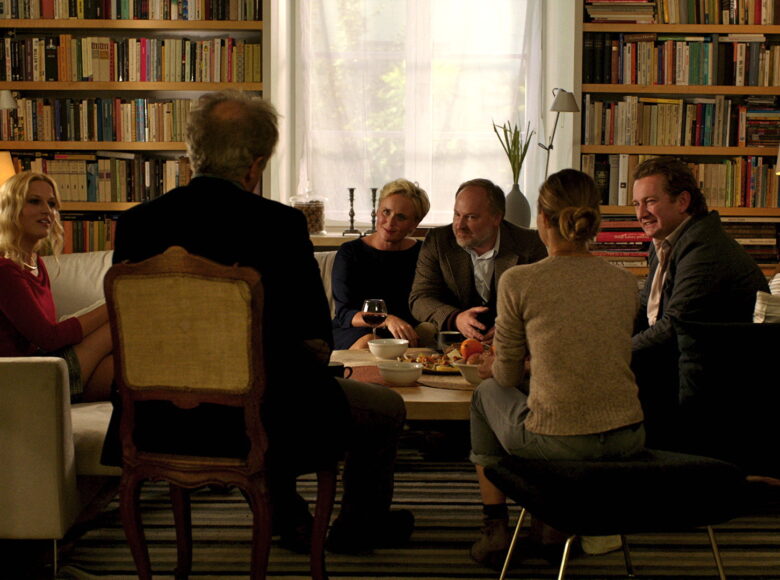 Kadr z filmu. Sześć osób – trzy kobiety i trzech mężczyzn siedzi w pokoju wokół stołu. Salon wypełniają między innymi regały pełne książek. Na stole naczynia, lampka wina. Wszystkie osoby patrzą na parę, która siedzi plecami do nas.