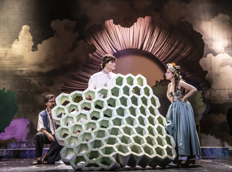 Na zdjęciu trójka osób. Na środku stoi młody mężczyzna ubrany w ludową koszulę z haftowanym kołnierzykiem, w rękach trzyma duży przedmiot w geometryczny wzór. Po prawej stronie stoi młoda kobieta z kwiatowym wiankiem na głowie. W tle duży okrąg-brama otoczona udrapowanym materiałem.