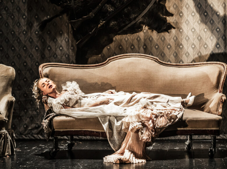 Na sofie w stylu XIX-wieku leży młoda kobieta ubrana w kremową suknię.