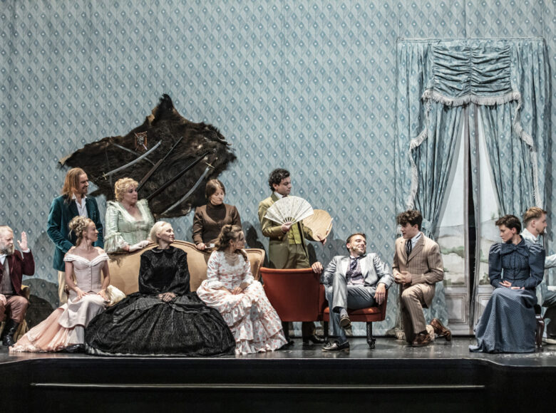 Scena zbiorowa Kobiety i mężczyzny w strojach z XIX wieku, siedzą i stoją w przestrzeni salonu, w tle ściana pokryta tapetą w błękitne romby oraz okno z udrapowanymi zasłonami.