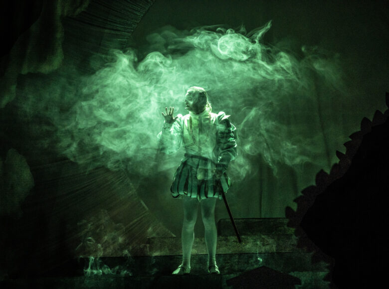 Mężczyzna w stroju XIX-wartownika, z pufiastymi spodniami. Stoi centralnie, oświetlonym zielonym światłem, dookoła unoszą się smugi dymu.