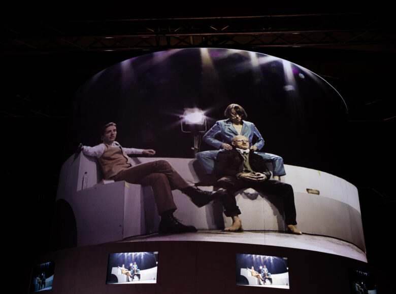 Ujęcie ekranów pokazujących zwielokrotniony ten sam obraz – dwóch mężczyzn i kobietę siedzących na białym podeście. Największy ekran na górze, częściowo wygięty w łuk, pod nim mniejsze ekrany.