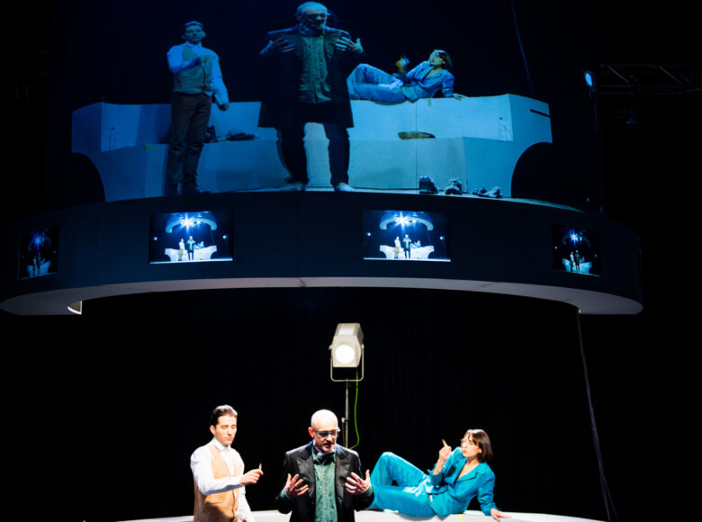 Obraz podzielony na dwie części w pionie. Na dole widać dwóch stojących mężczyzn i kobietę na wpół leżącą na białym postumencie. Ponad nimi ten sam obraz wyświetlony jest na dużym ekranie.