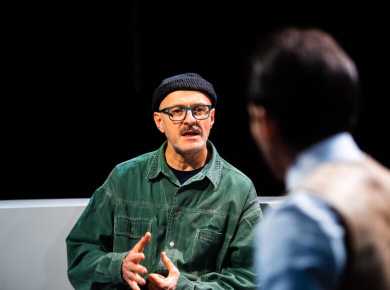 Na zdjęciu dwie osoby. Przodem do kadru mężczyzna w okularach i czarnej wełnianej czapce na głowie, wydaje się mówić. Na pierwszym planie niewyraźna sylwetka osoby odwróconej plecami.