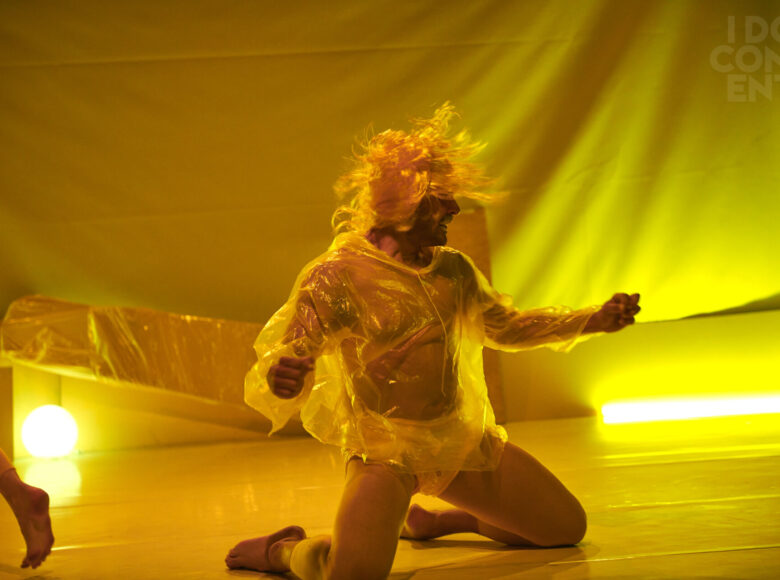 Osoba ubrana w foliowy płaszcz przeciwdeszczowy i złotą perukę klęczy na scenie. Ręce ma uniesione na boki. Scenę oświetla żółte światło.