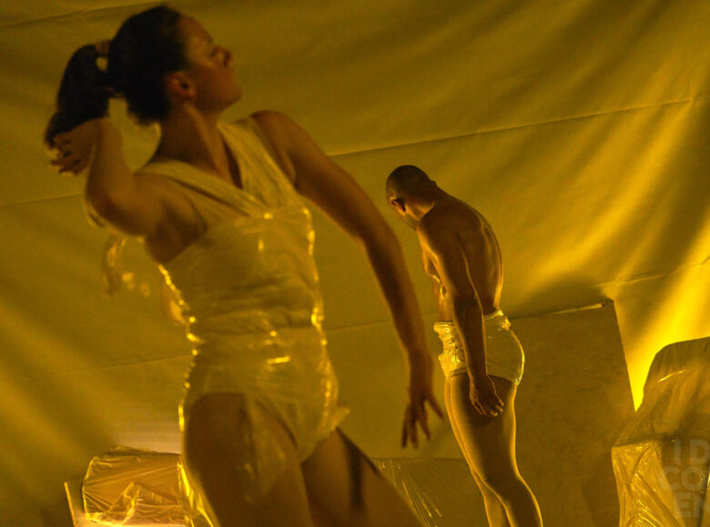 Na pierwszym planie tańcząca kobieta. Tułów owinięty ma przeźroczystą folią. Za nią w tle stoi półnagi mężczyzna.