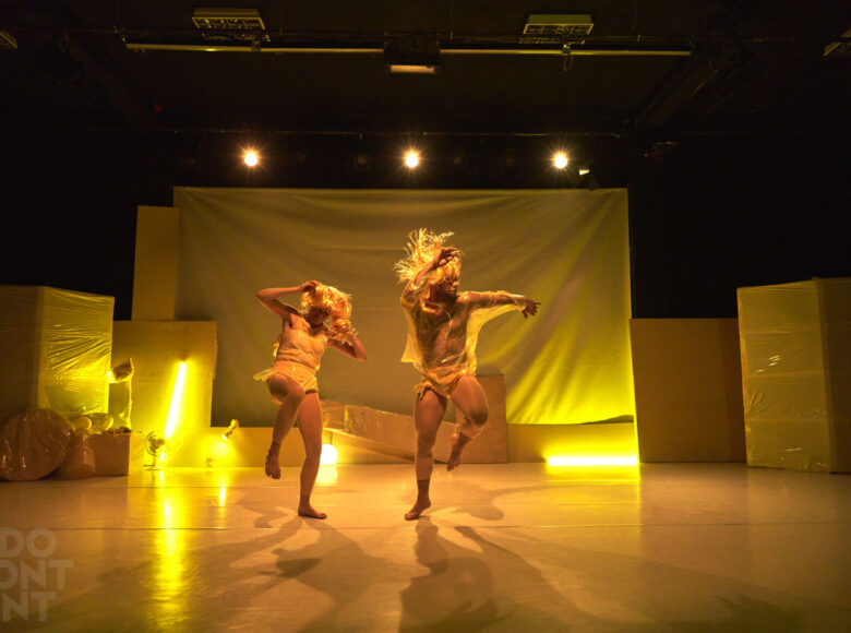 Kolorowa fotografia. Na scenie dwie osoby ubrane w foliowe płaszcze przeciwdeszczowe i złote peruki. Wykonują chaotyczne podskoki. Za nimi w tle scenografia składająca się z wielu, różnej wielkości zafoliowanych pudeł. Scena oświetlona jest żółtym światłem.