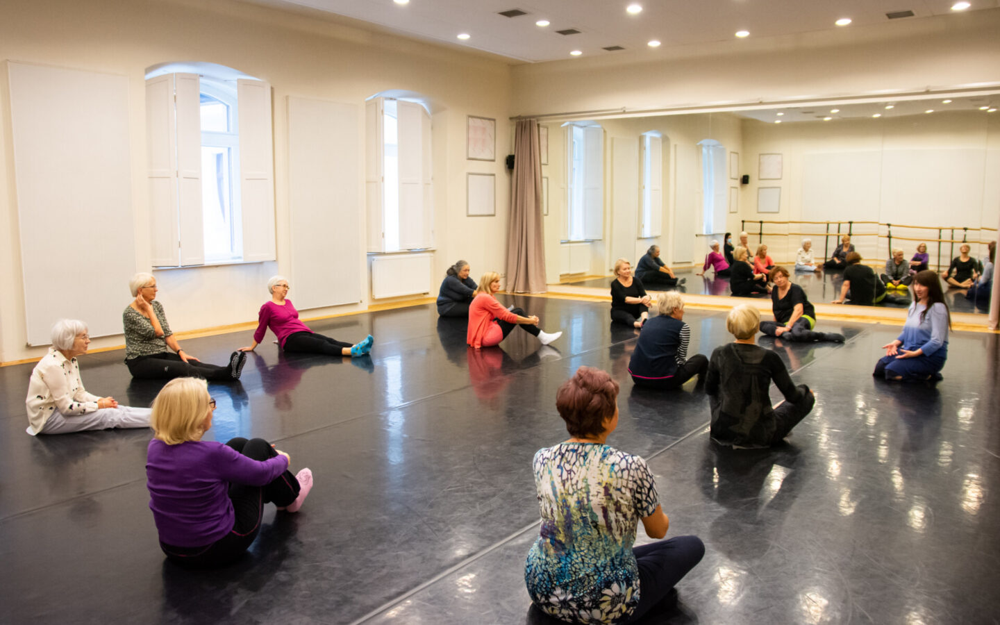 Kolorowa fotografia. Wnętrze sali prób. Na tanecznej podłodze siedzi w rozsypce grupa starszych kobiet ubranych w sportowe stroje. Patrzą i słuchają prowadzącej zajęcia młodszej kobiety.