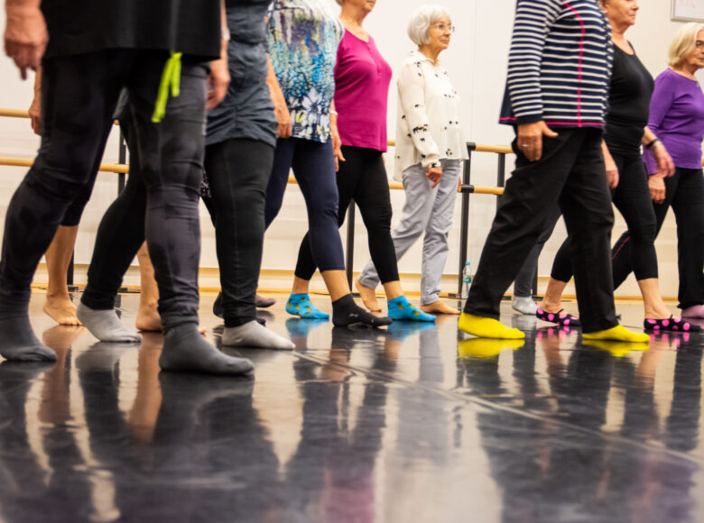 Kolorowa fotografia. Ujęcie z dołu przedstawiające podłogę taneczną, po której kroczy grupa starszych kobiet ubranych w sportowe stroje.
