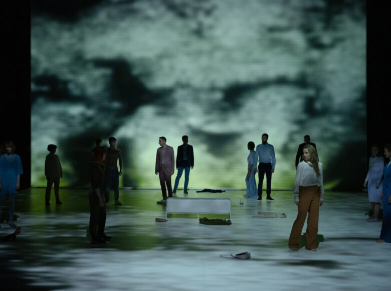 Scena grupowa. Grupa ludzi stoi rozproszona na przestrzeni oświetlonej wizualizacjami w kolorystyce szarości i zieleni.
