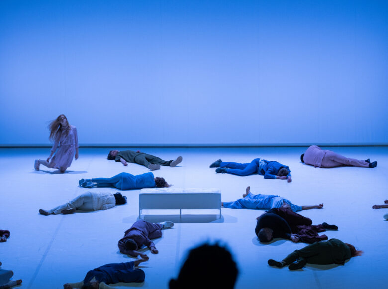 Scena zbiorowa skąpana w niebieskim świetle.. Grupa ludzi leży rozproszona na białej podłodze. Jedynie w lewym górnym rogu jedna kobieta w ruchu, wydaje się przewracać. Centralnie na środku stoi biała, prostokątna sofa.