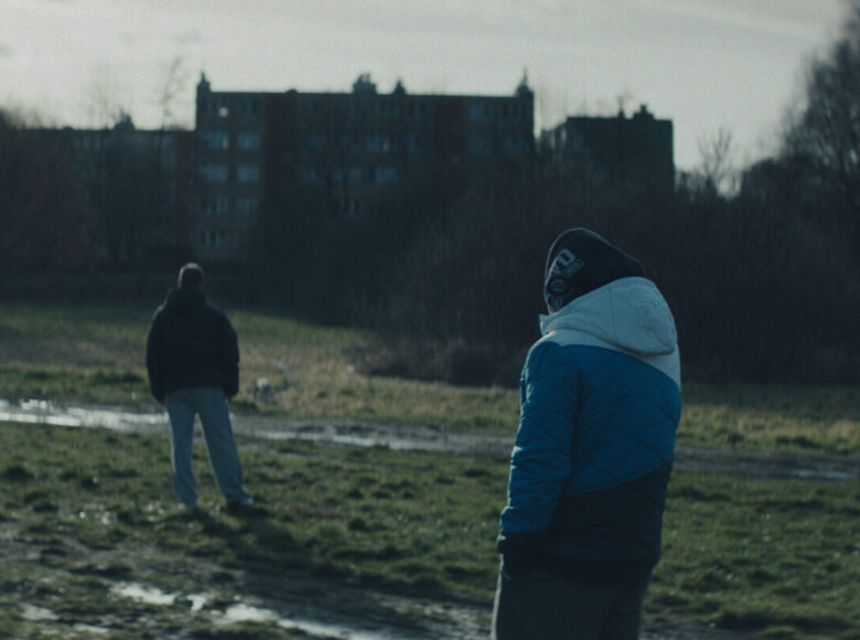 Kadr z filmu. Dwóch mężczyzn stoi na zewnątrz. Odwróceni plecami do nas, ubrani w dresy, kurtki. Jeden stoi kilka metrów przed drugim. Stoją na trawie, w której są koleiny i kałuże. Przed nimi bloki, bezlistne drzewa. Tło jest rozmyte.