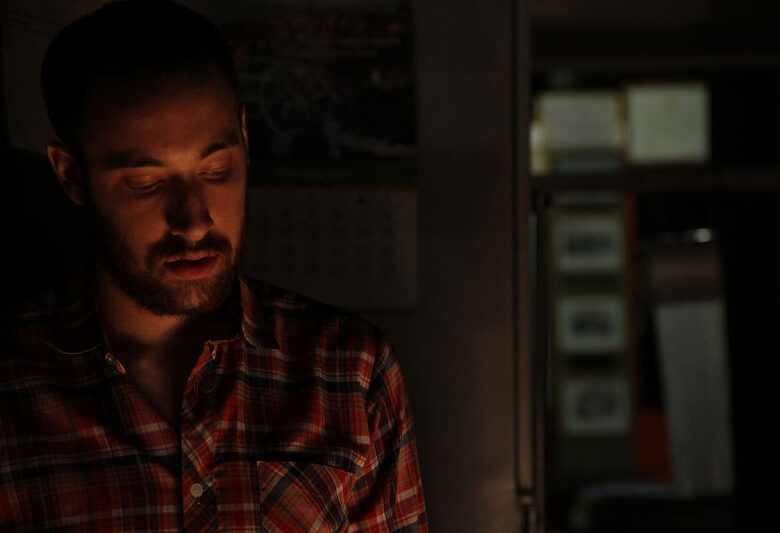 Kadr z filmu. Młody mężczyzna w koszuli w kratę. Ma ciemny zarost, opuszczony wzrok. Stoi w ciemnym wnętrzu, oświetlony od dołu.