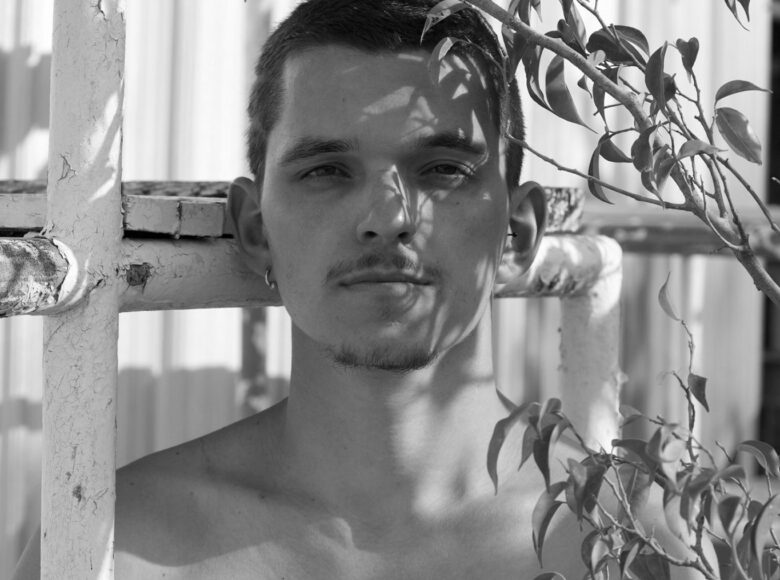 Biało-czarne zdjęcie. Portret młodego mężczyzny. Mężczyzna nie ma na sobie koszuli. Piera się o drewnianą barierkę, obok niego rośnie małe drzewko.