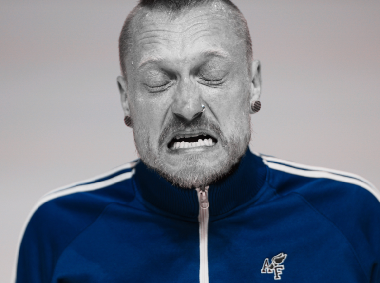 Czarno-białe, artystyczne zdjęcie. Portret mężczyzny. Ma zamknięte oczy i otworzone usta. Po czole spływa mu pot. Mężczyzna ma na sobie błękitna bluzę na zamek. Bluza jest jedynym kolorowym elementem zdjęcia. Reszta jest czarno-biała.