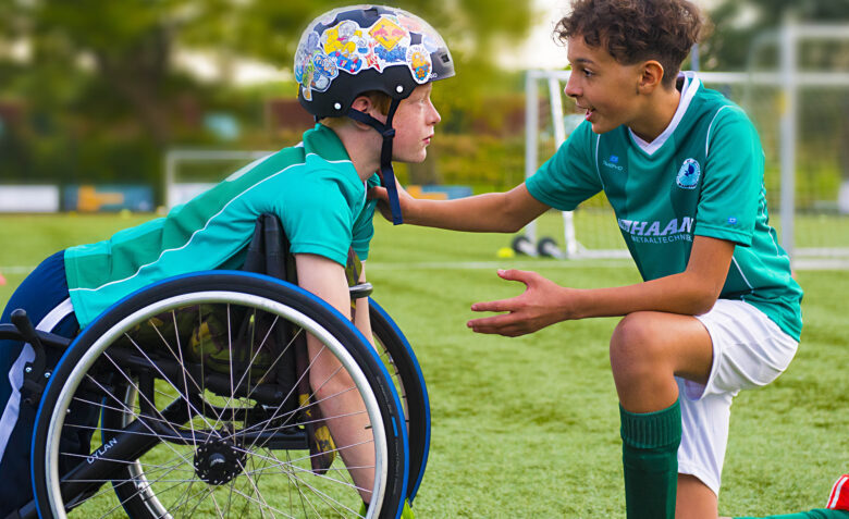 Na zdjęciu dwaj chłopcy na boisku piłkarskim – jeden z nich oparty jest na wózku inwalidzkim, jest w kasku – drugi przyklęka przed nim i klepie po ramieniu, tłumaczy i gestykuluje.