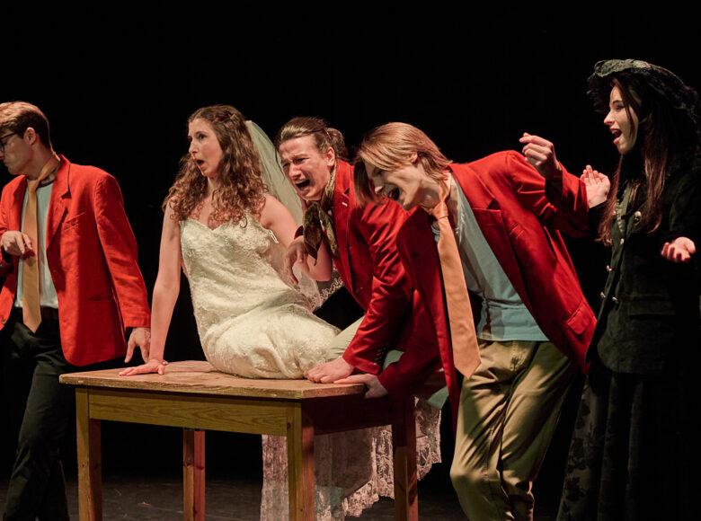 Zdjęcie ze spektaklu. Na scenie znajduje się stół, na którym siedzi dziewczyna w sukni ślubnej i z welonem na głowie. Wokół niej stoją chłopcy w szkolnych mundurkach: czerwonych marynarkach i pomarańczowych krawatach. Obok jednego z chłopców stoi dziewczyna w czarnej koronkowej sukni i kapeluszu na głowie.