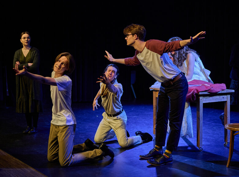 Zdjęcie ze spektaklu. Na scenę pada niebieskie światło. Na stole siedzi zgarbiona dziewczyna w sukni ślubnej. Przed nia kleczą dwaj chłopcy i wyciągają przed siebie ręce, patrzą w stronę publiczności. Trzeci stoi, rozkłada obie ręce w bok i lekko pochyla się do przodu.