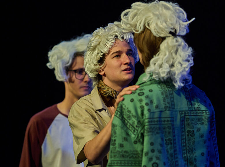 Zdjęcie ze spektaklu. Dwóch chłopaków w siwych perukach na głowach stoi na przeciwko siebie. rozmawiają ze sobą. Za nimi stoi trzeci chłopak w peruce i patrzy przed siebie.