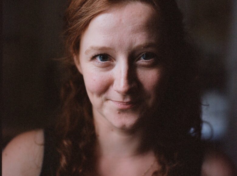 Zdjęcie. Portret młodej kobiety. Ma rude, kręcone włosy i srebrny łańcuszek na szyi. Patrzy przed siebie, uśmiecha się.