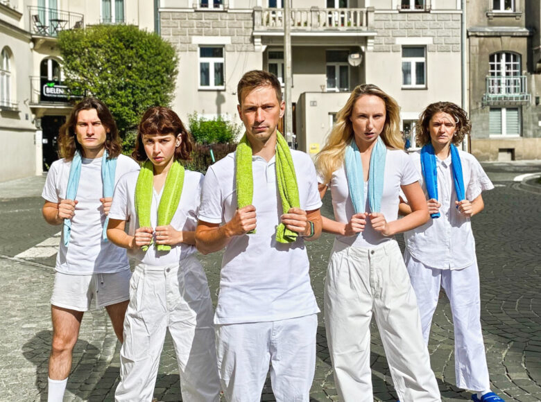 Wybrukowana ulica, w tle kamienice. Po środku ulicy stoi pięć osób. Wszyscy ubrani są w białe spodnie i koszulki. Na szyję mają zarzucone kolorowe ręczniki, które trzymają w dłoniach.