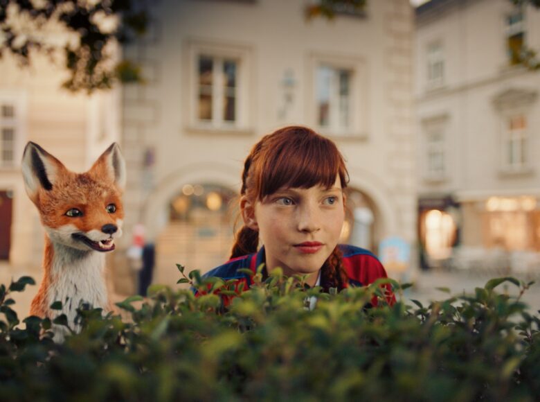 Na zdjęciu dziewczyna razem z animowanym lisem wyglądający zza żywopłotu, w tle widok na uliczkę i kamienice.
