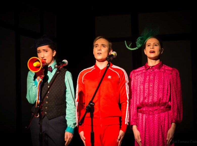 W szeregu przed mikrofonami stoją trzy osoby. Od lewej : kobieta z zabawkowym megafonem, mężczyzna w czerwonym dresie oraz kobieta w jaskrawej różowej sukience w czarne kropki. Wszyscy stoją na baczność oraz śpiewają.