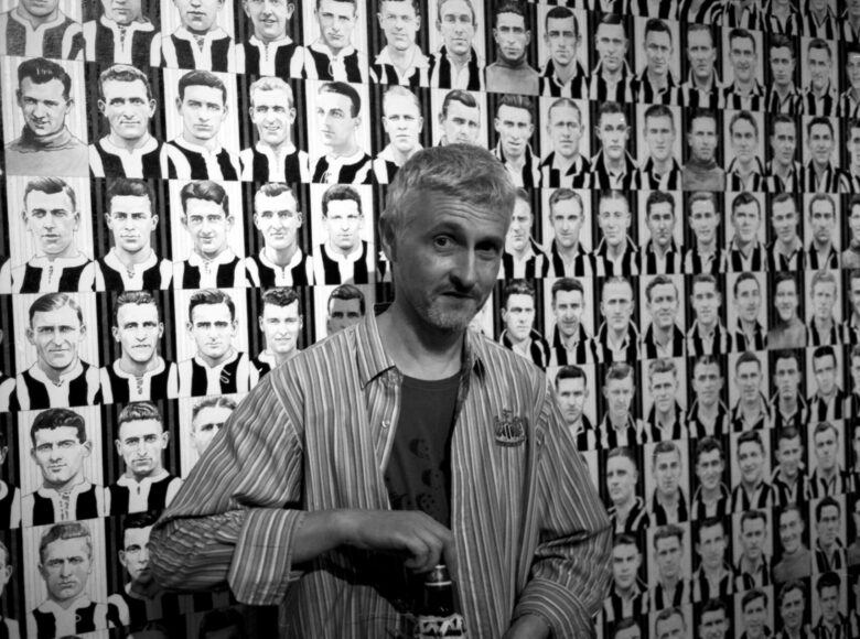 Grafika na ścianie. Składa się z namalowanych setek małych popiersi piłkarzy grupy Niukasl Junajted, którzy mają koszulki w biało-czarne pionowe pasy. Przed ściana soi mężczyzna w koszuli w paski. Patrzy przed siebie i otwiera butelkę piwa.